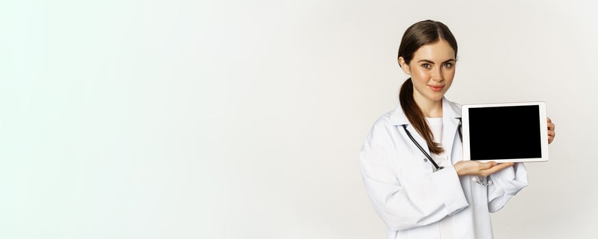 Online medical help, remote appointment. Smiling beautiful woman doctor showing digital tablet screen, demonstrating website logo, standing over white background photo