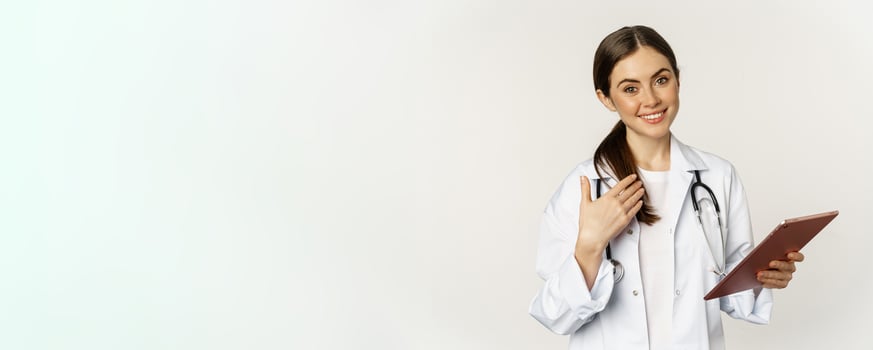 Smiling brunette female doctor, holding digital tablet, pointing at herself with friendly face, standing in white coat over white background photo