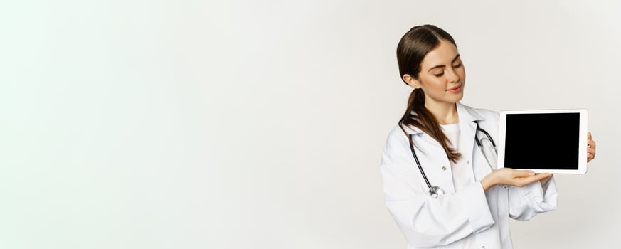 Image of woman doctor, female healthcare worker showing online medical website, digital tablet screen and smiling, standing in white coat over white background photo
