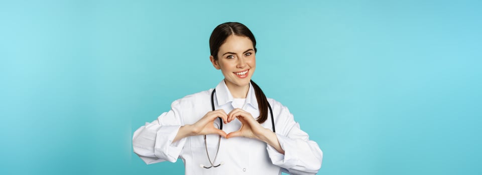 Young doctor, woman physician showing heart, love sign and smiling, care for patients in clinic, standing in white coat over blue background photo
