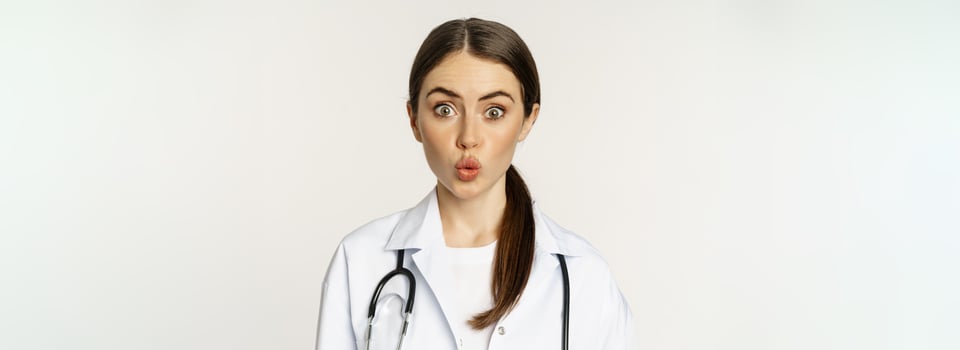 Portrait of woman doctor looking surprised, amazed, reaction of interest and amusement, standing in hospital uniform over white background photo