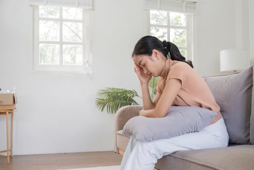 Woman Sitting on Couch Holding Head in Pain, Suffering from Headache and Feeling Sick in Bright Living Room photo