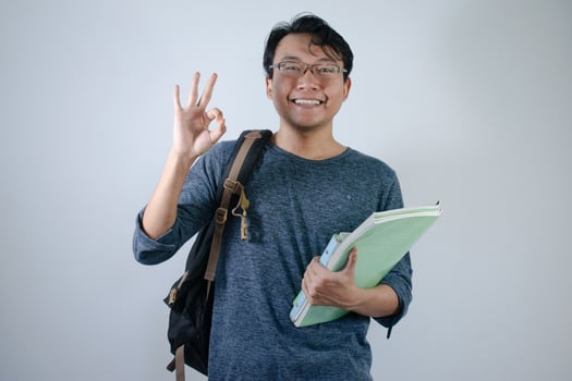 Asian young male student wearing bag and holding book smiling and happy expression. Ok hand sign gesture with isolated white background photo
