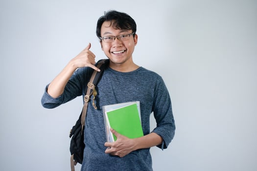 Asian young male student wearing bag and carrying books smiling and happy expression. Call me hand sign gesture with isolated white background photo