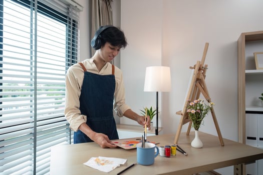 Young Artist Painting at Home Studio with Headphones, Wearing Apron, and Surrounded by Art Supplies and Natural Light photo