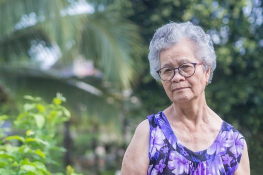 ortrait of an elderly Asian woman with short gray hair, wearing glasses, smiling, and looking at the camera while standing in a garden. photo