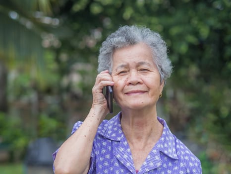 Happy beautiful senior woman is using a smartphone, smiling and looking at the camera while standing in a garden. Space for text. Aged people and communication concept photo
