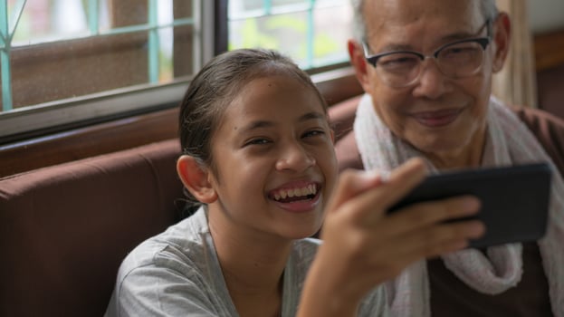 Cheerful adorable Thai girl holding mobile phone enjoy taking selfie photo with her senior grandfather in living room. Using call with smartphone. Bonding relationship in family.