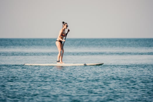Sea woman sup. Silhouette of happy middle aged tanned woman in rainbow bikini, surfing on SUP board, confident paddling through water surface. Idyllic sunset. Active lifestyle at sea or river. photo