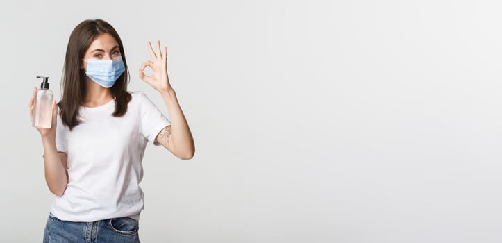 Covid-19, health and social distancing concept. Portrait of smiling brunette girl in medical mask, showing hand sanitizer and okay gesture photo