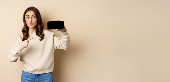 Image of girl pointing finger, showing mobile phone horizontal screen and looking amazed by application, standing over beige background photo