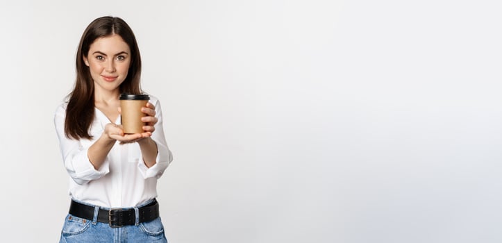 Image of modern woman, office lady holding takeaway coffee cup and smiling, standing over white background photo