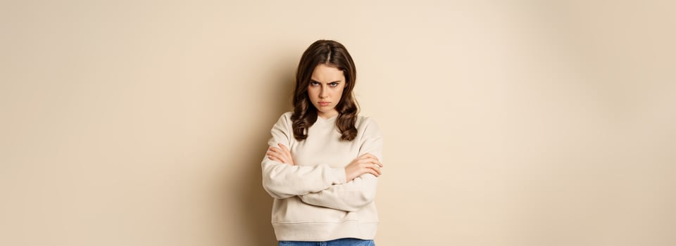 Angry girlfriend sulking, cross arms on chest and look offended, staring moody at camera, standing over beige background photo