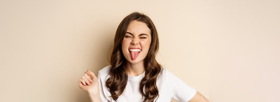 Close up of happy, carefree beautiful woman posing silly, showing tongue, having fun, standing in casual white t-shirt against beige background photo