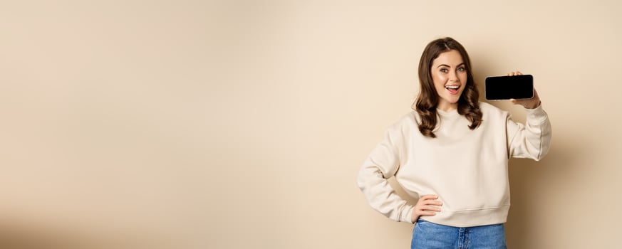 Smiling beautiful female model demonstrating horizontal smartphone screen, mobile application, standing over beige background photo