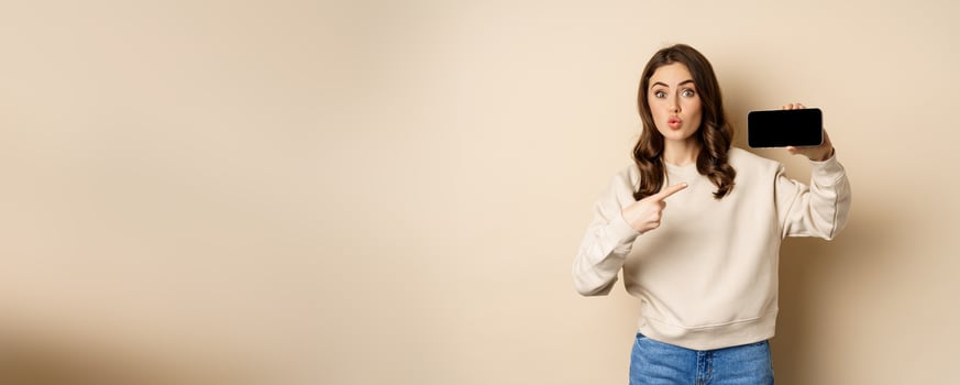 Image of girl pointing finger, showing mobile phone horizontal screen and looking amazed by application, standing over beige background photo
