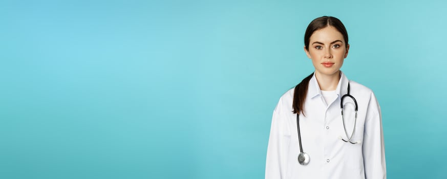 Medical staff and doctors concept. Young smiling female doctor, healthcare worker in white coat and stethoscope, looking confident, waiting patients, blue background photo