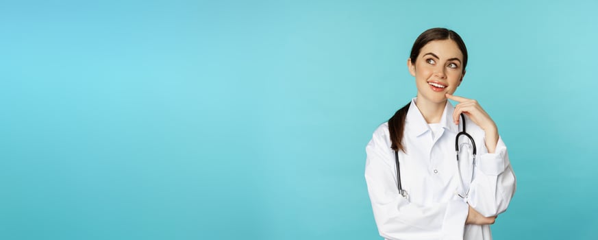 Young female doctor, hospital worker in white coat, thinking and looking away thoughtful, searching solution, standing over toquoise background photo