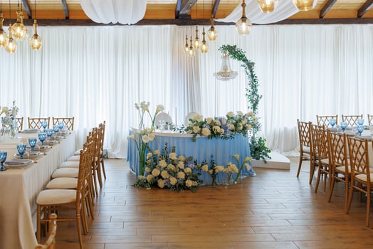 A large room with a blue table and white curtains. The room is decorated with flowers and has a blue table with white flowers on it. There are many chairs around the table photo