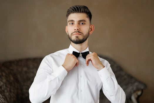 A man in a white shirt and black bow tie is getting ready to go out. He is standing in front of a brown wall photo