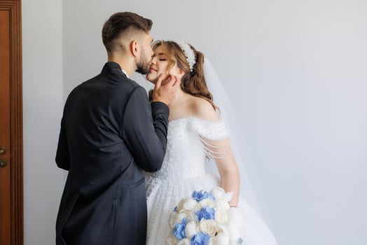 A man and woman are kissing in front of a white door. The man is wearing a suit and tie, and the woman is wearing a wedding dress. Scene is romantic and intimate photo