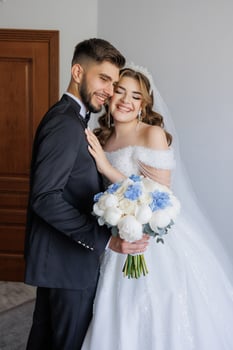A bride and groom are posing for a picture with a bouquet of flowers. The bride is wearing a white dress and the groom is wearing a suit. Scene is happy and celebratory photo