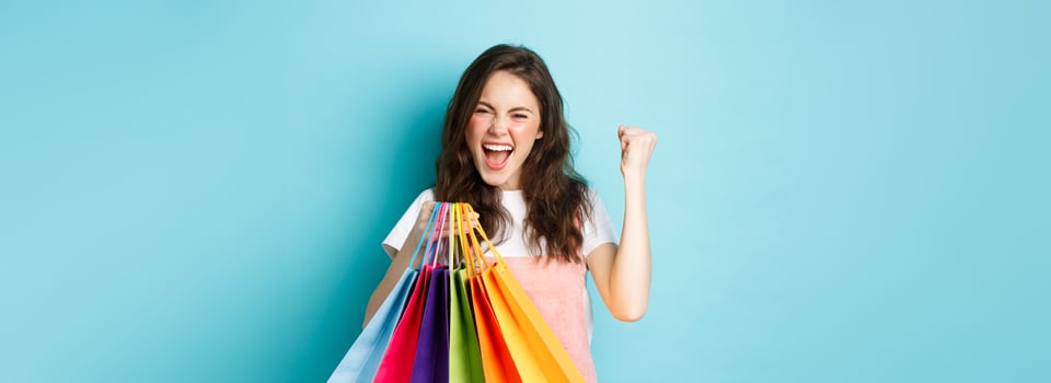 Excited young woman scream of joy, say yes and make fist pump satisfied, buying gifts with discounts and triumphing, holding shopping bags, standing over blue background photo