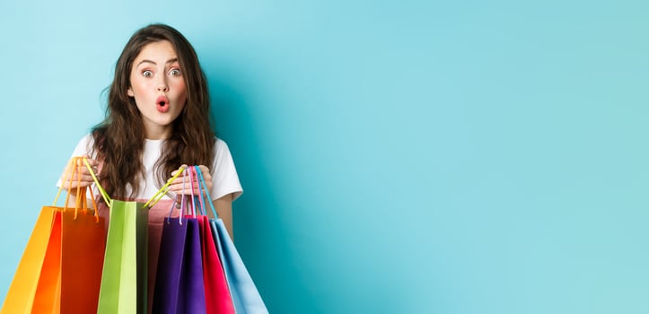 Amazed young woman shopaholic, holding colorful shopping bags and look amused at next shop, buying things in store, standing over blue background photo