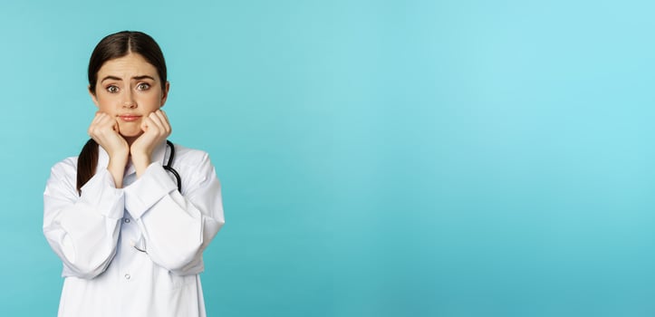 Scared and worried woman doctor in white coat, looking anxious and insecure, shaking from fear, standing over torquoise background photo