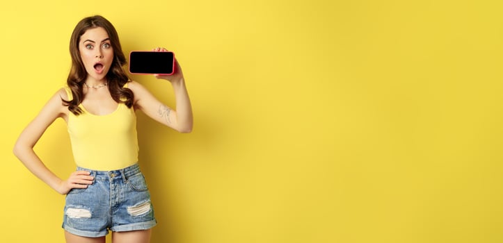 Shocked and amazed stylish girl, showing smartphone screen, app on horizontal mobile phone display, standing in summer clothes over yellow background photo
