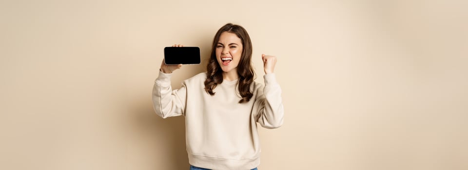 Happy girl showing mobile phone horizontal screen and celebrating, chanting with joyful face, standing over beige background photo