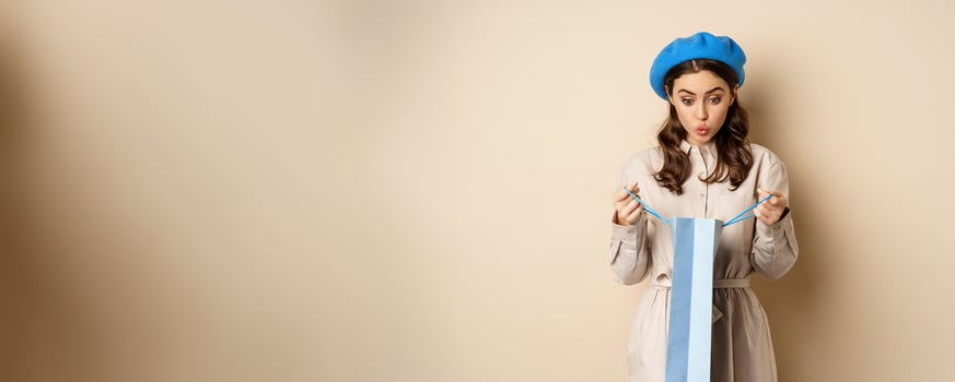 Image of girl looking surprised after opening gift bag, looking at present amazed, standing in stylish outfit over beige background photo
