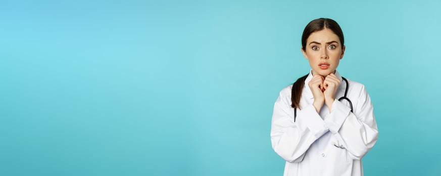 Worried and concerned physician, hospital worker in white lab coat, looking scared and nervous at camera, standing with stethoscope over torquoise background photo