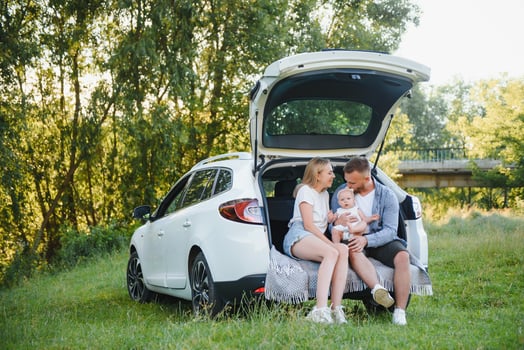 Pretty young married couple and their daughter are resting in the nature. The woman and girl are sitting on open car boot. The man is standing near them. They are smiling photo