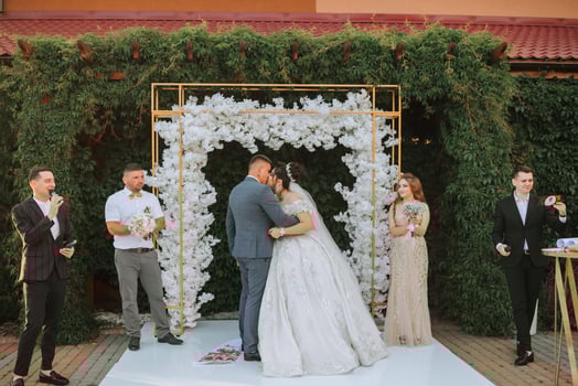 A bride and groom are kissing under a white arch. The bride is wearing a long white dress and the groom is wearing a suit. There are several other people in the background photo