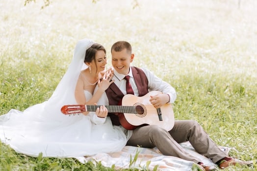 A couple is sitting on a blanket in a grassy field, with a guitar in front of them. The man is wearing a red vest and the woman is wearing a white wedding dress photo