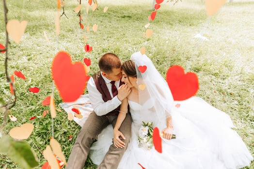 A bride and groom are sitting on a blanket in a field with red hearts surrounding them. The scene is romantic and intimate, with the couple embracing each other photo