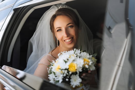 A bride is sitting in a car with a bouquet of flowers. She is smiling and looking out the window photo