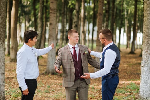 Three men are standing in a forest, one of them is wearing a suit and tie. They are all looking at something, possibly a camera or a piece of equipment. Scene is casual and relaxed photo