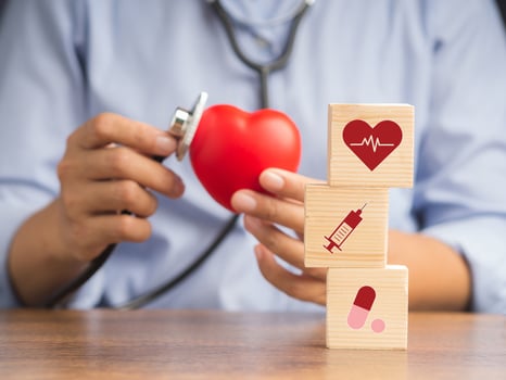 Hand of a doctor holding a stethoscope checking a red heart shape and wooden blocks with healthcare medical icons while sitting in the hospital or clinic. Medical, treatment, and healthcare concept photo