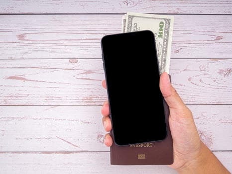 Close-up of hand holding a smartphone with passport and US dollars banknote on wooden table background. Communication and travel concept photo