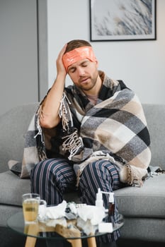 Sick adult man who has bad cold or seasonal flu virus is fighting illness at home. Unhappy middle aged man is sitting on sofa wrapped in warm scarf and plaid, holding paper tissue and sneezing. photo