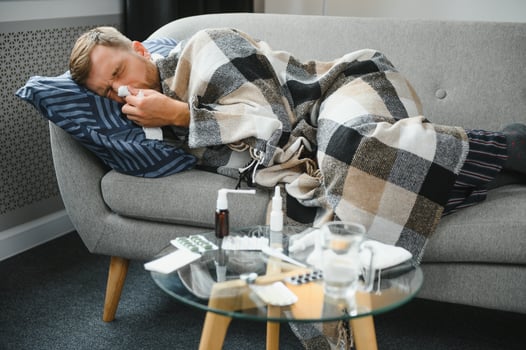 Sick bearded man who has bad cold or seasonal flu sitting on couch at home. Guy with fever wearing warm plaid shivering with worried face expression. photo