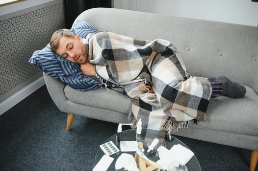 Sick bearded man who has bad cold or seasonal flu sitting on couch at home. Guy with fever wearing warm plaid shivering with worried face expression. photo