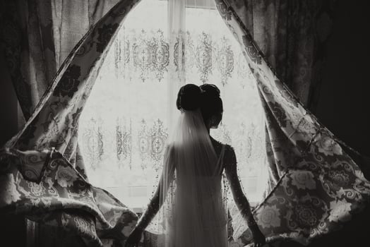 A woman in a wedding dress stands in front of a window with a curtain open. Concept of anticipation and excitement for the upcoming wedding photo