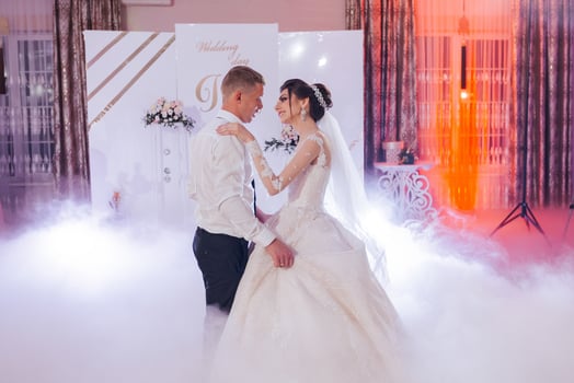 A bride and groom are dancing in a foggy room. The bride is wearing a white dress and the groom is wearing a white shirt photo