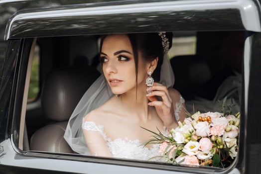 A woman in a wedding dress is sitting in a car with a bouquet of flowers. She is wearing a tiara and earrings photo