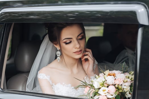 A woman in a wedding dress sits in a car with a bouquet of flowers. She is wearing a veil and has her hands on her head. The car is a black SUV photo