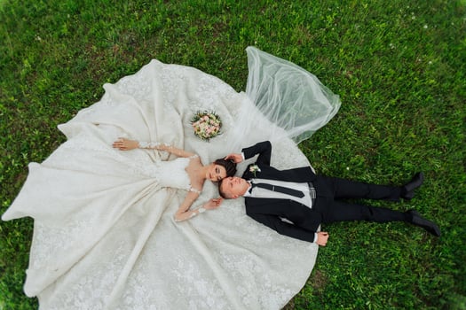 A bride and groom are lying on the grass, with the bride wearing a white dress. The bride is holding a bouquet of flowers in her lap. The scene is romantic and intimate, capturing the love photo