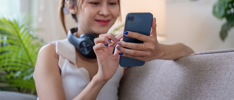 Young Woman Using Smartphone at Home with Headphones Around Neck, Relaxing on Couch in Modern Living Room photo
